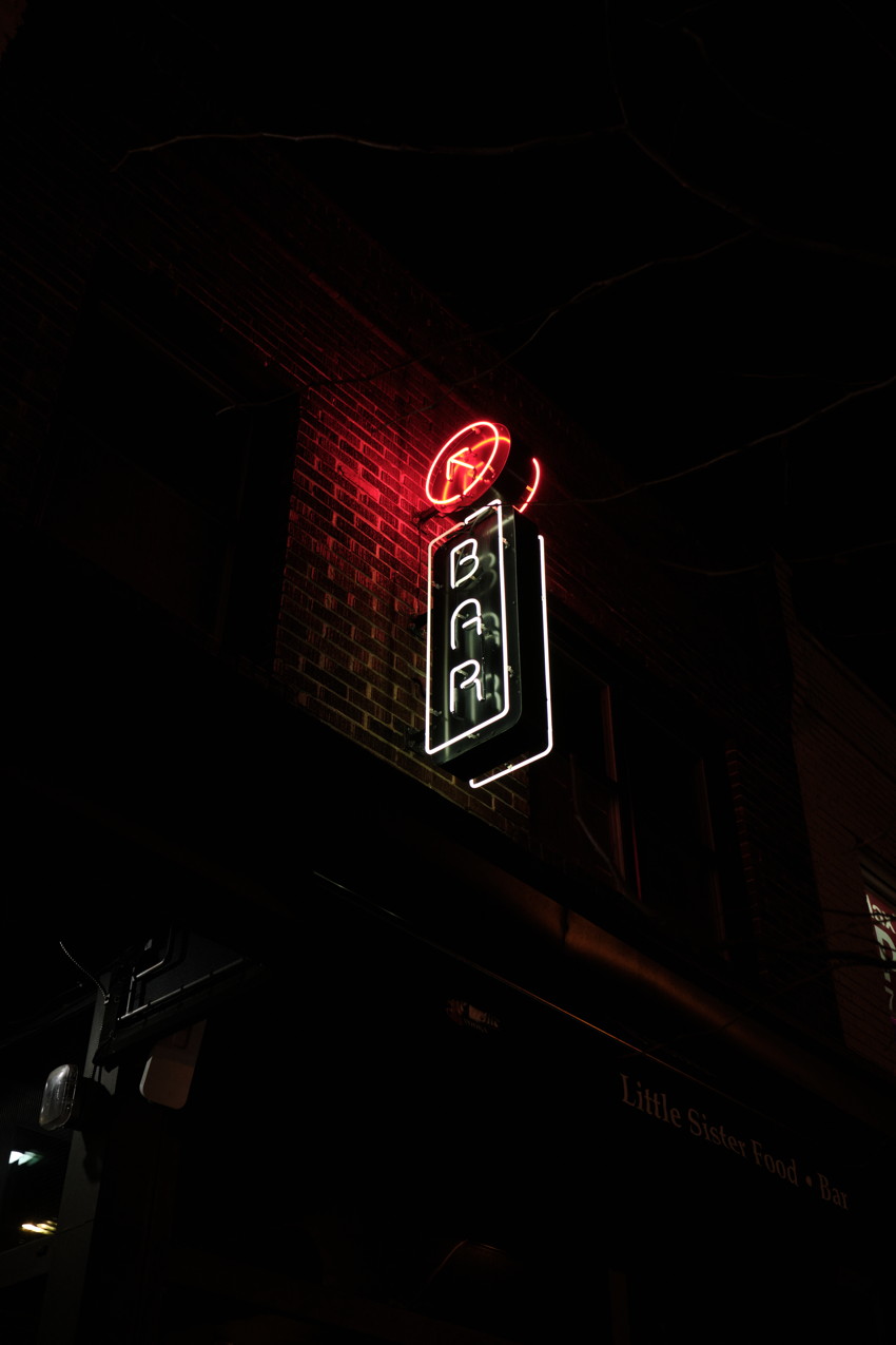 Vertical neon 'BAR' sign in red and white mounted on dark brick building at night, angled upward.