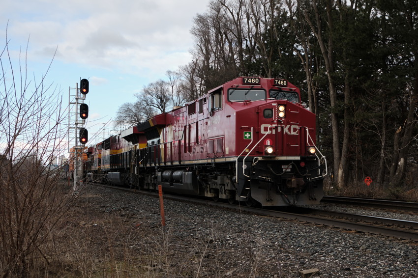 Red freight locomotive (7460) on tracks at right, signal lights left, gravel foreground, leafless trees background.