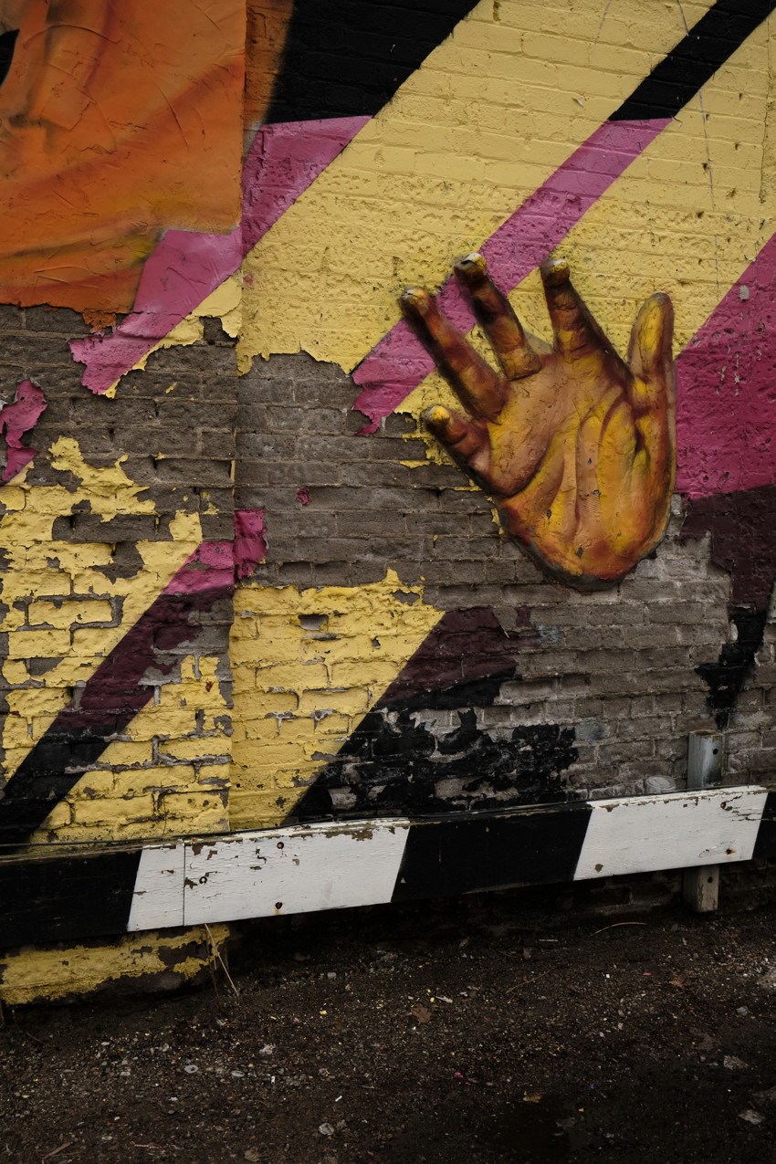 Peeling brick wall mural with diagonal yellow and pink stripes and a painted yellow hand; black-and-white barrier at base.
