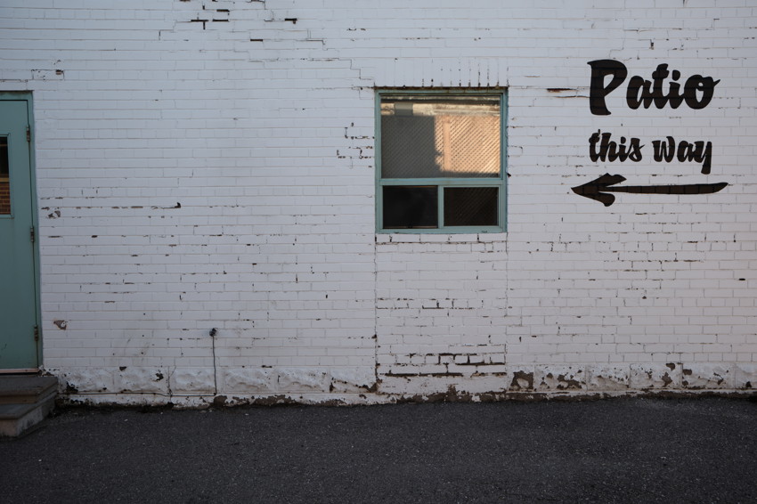 White brick wall, small green-framed window, teal door at left, painted 'Patio this way' with arrow.