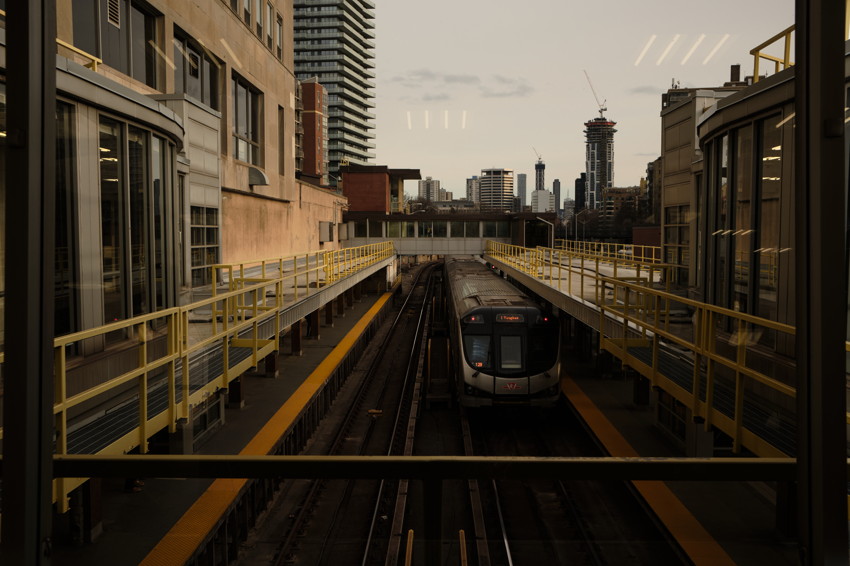 Centered commuter train arriving on elevated tracks between yellow-railed platforms, urban skyline in background.