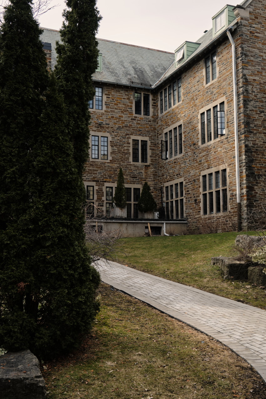 Stone multi-story building with many windows; paved walkway from foreground, evergreen trees left, grassy lawn.