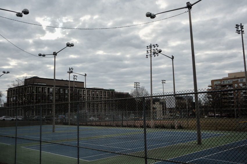 Chain-link fence, empty blue-green tennis courts, tall light poles, overcast sky, mid-rise buildings.