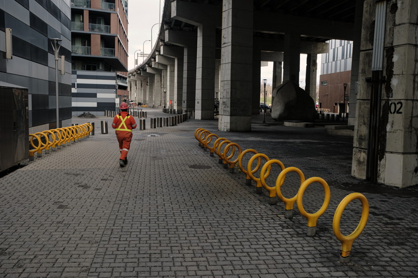 Worker in orange walking under concrete overpass; curved row of yellow circular bike racks along paved plaza.
