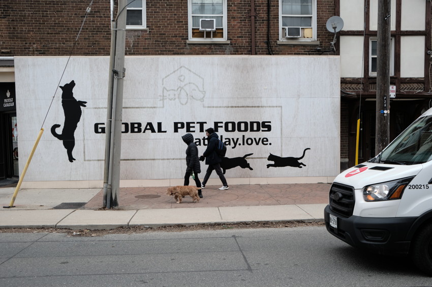 Two people walking a small dog past mural reading 'GLOBAL PET FOODS' with black animal silhouettes.