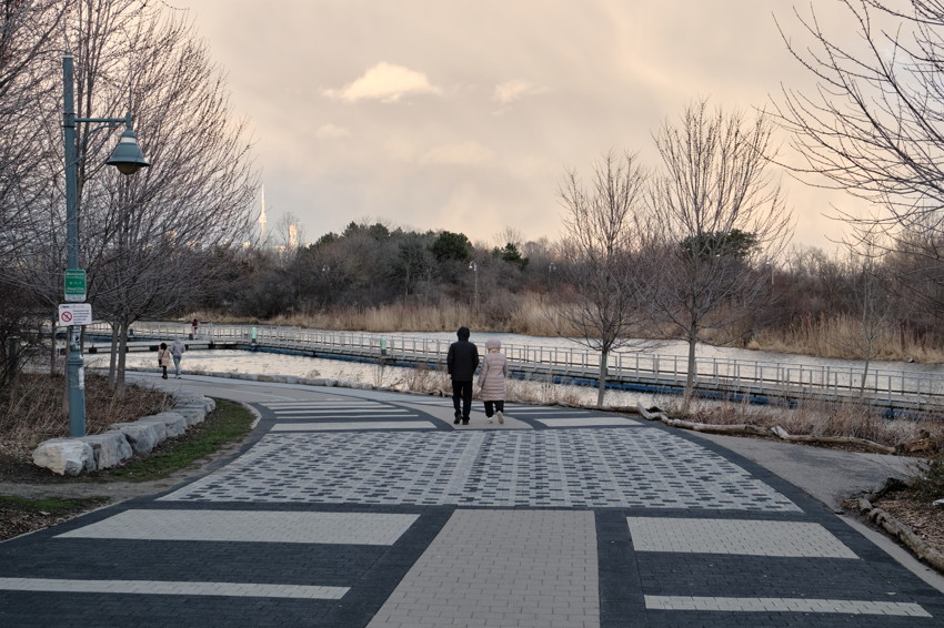 Paved promenade with geometric tiles, two people walking away toward boardwalk, bare trees and river.