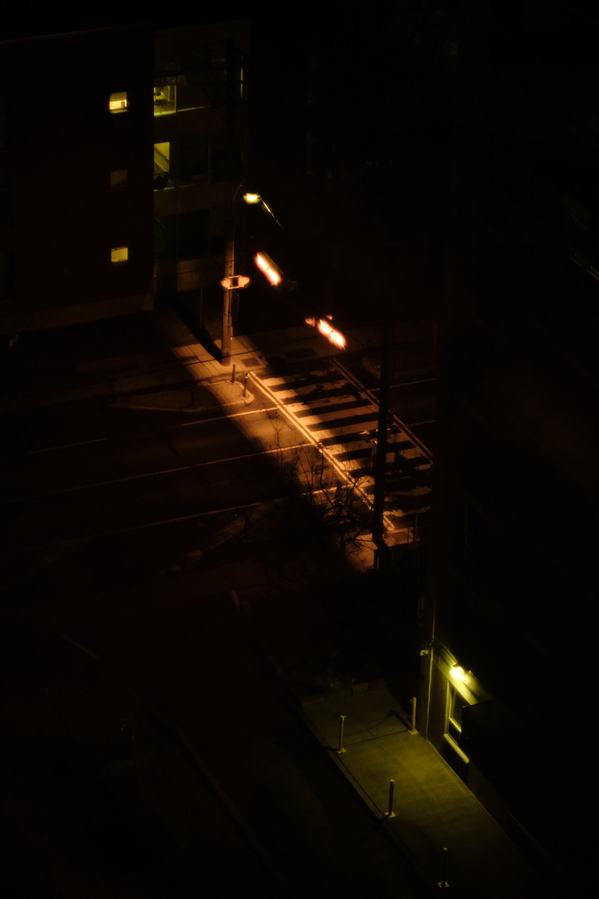 High-angle nighttime view of an empty crosswalk lit by orange streetlights, adjacent dark buildings and a lit doorway.