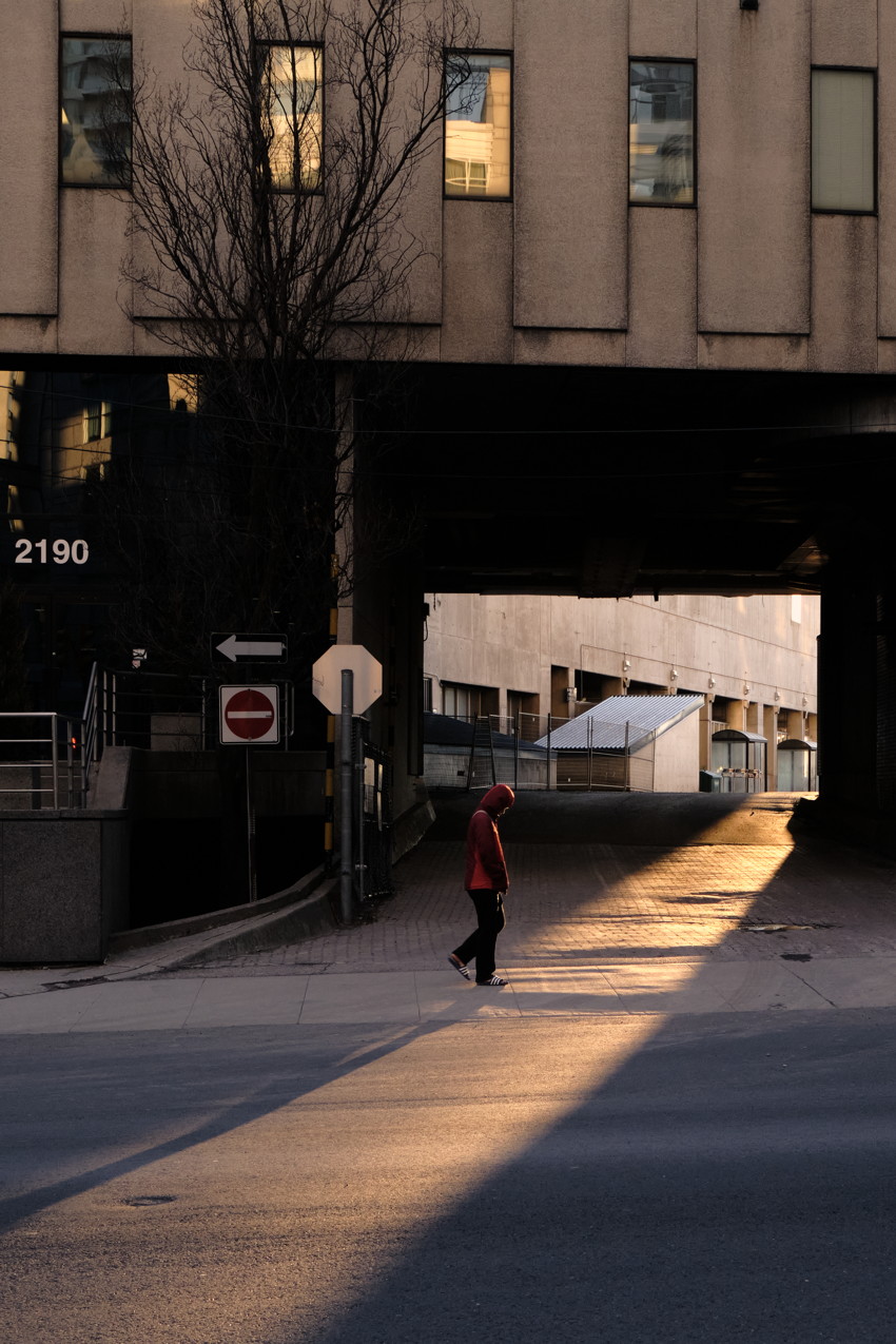 Person in red hoodie walking through sunlit diagonal beam under a concrete building overpass.