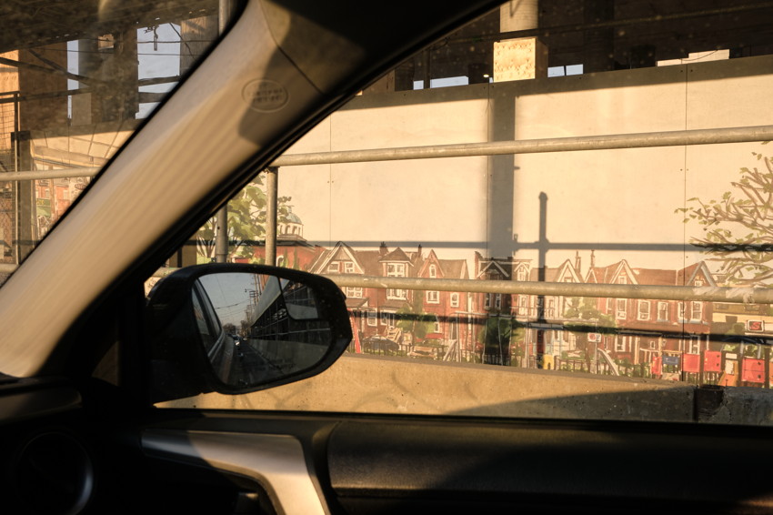 Car interior view through passenger window and mirror; metal railing and mural of row houses on wall outside.