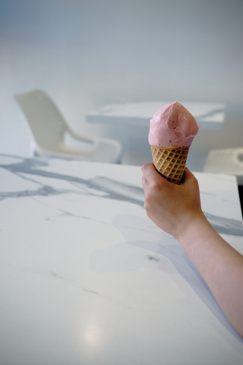 Child's hand holding waffle cone with pink ice cream over white marble table; blurred plastic chairs in background.