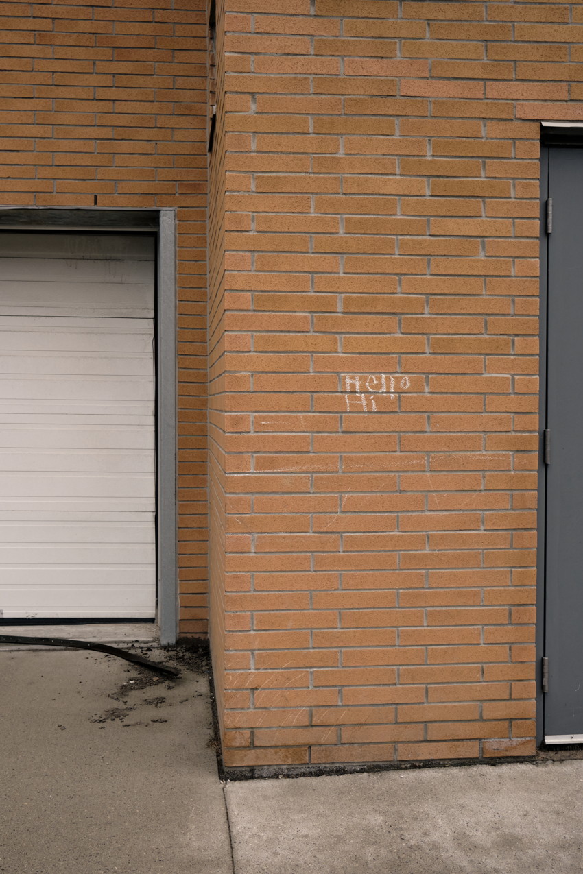 Tan brick building corner with white roll-up garage door left, gray door right, small chalk "hello" on bricks.