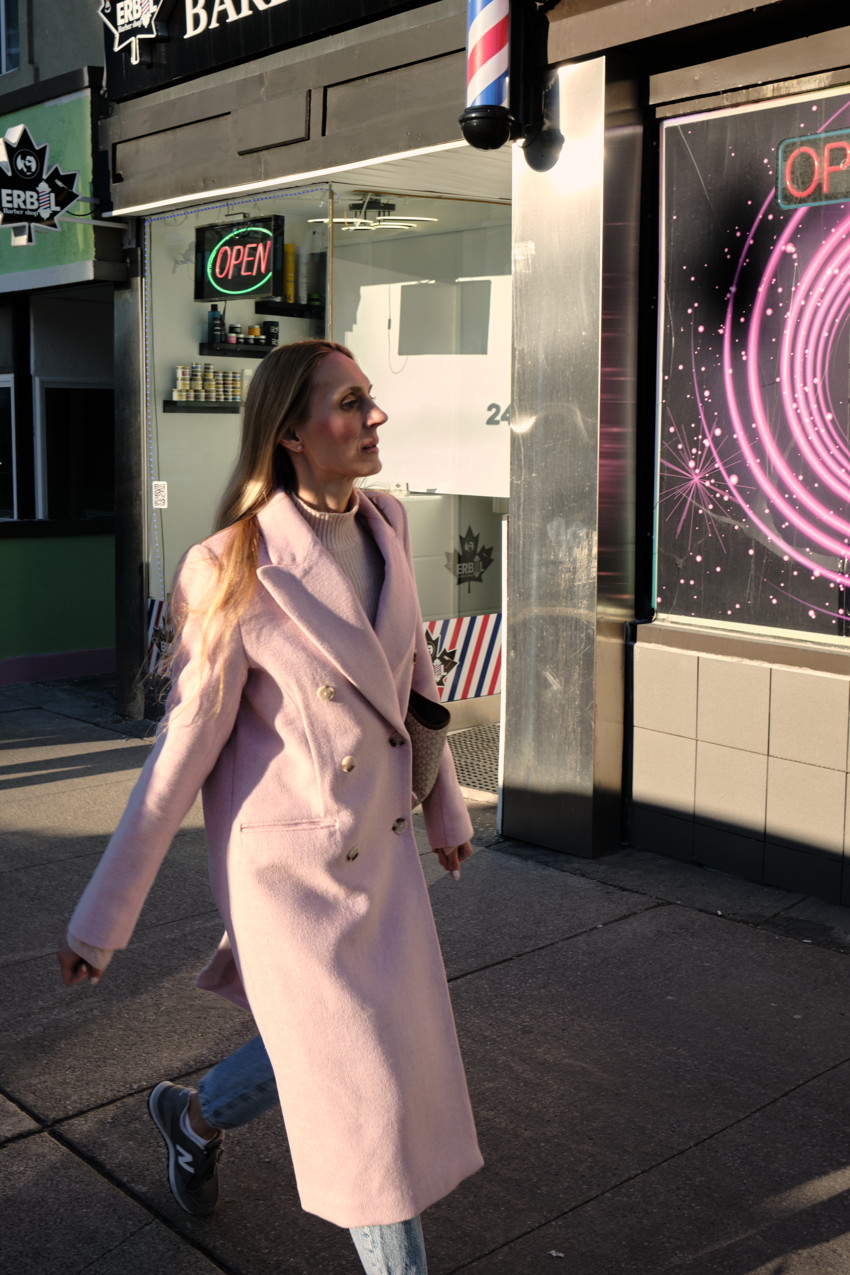 Person in long pink coat walking past barbershop storefront with barber pole and neon OPEN sign.