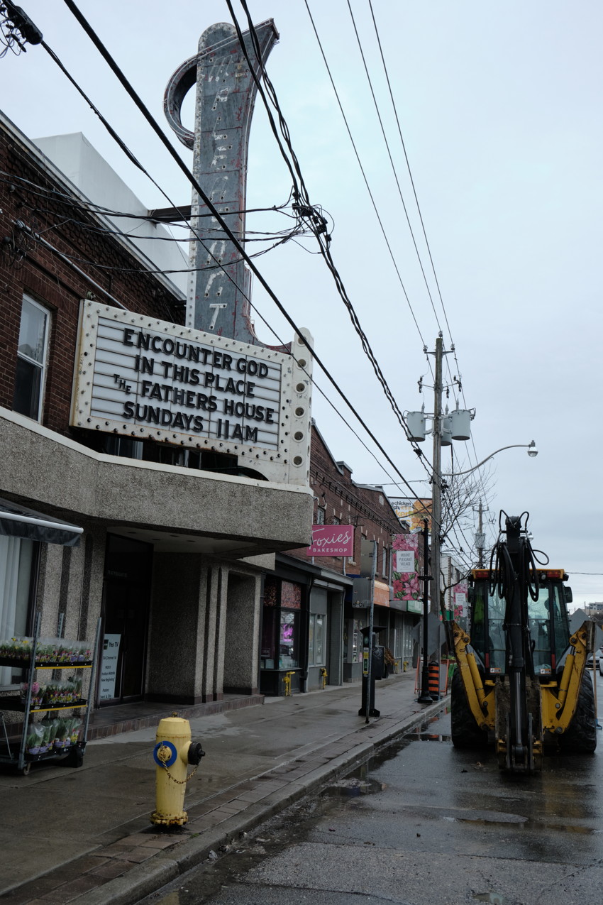 Street view with vintage theater marquee 'ENCOUNTER GOD IN THIS PLACE', storefronts, fire hydrant, parked backhoe.