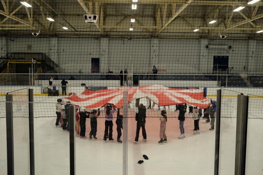 Indoor ice rink through safety netting; skaters form circle holding large red-and-white striped fabric.