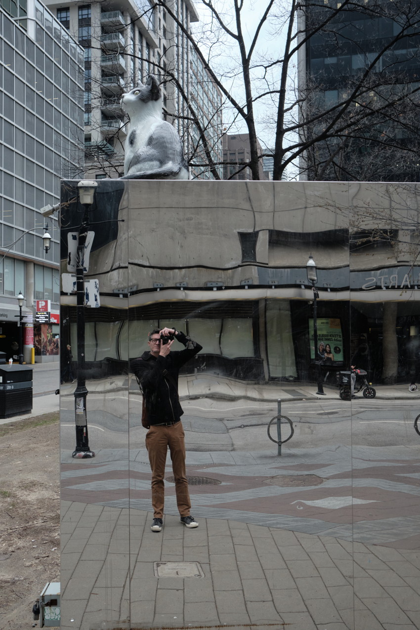 Person photographing their reflection in a distorted mirrored wall beneath a large cat sculpture in a city plaza.
