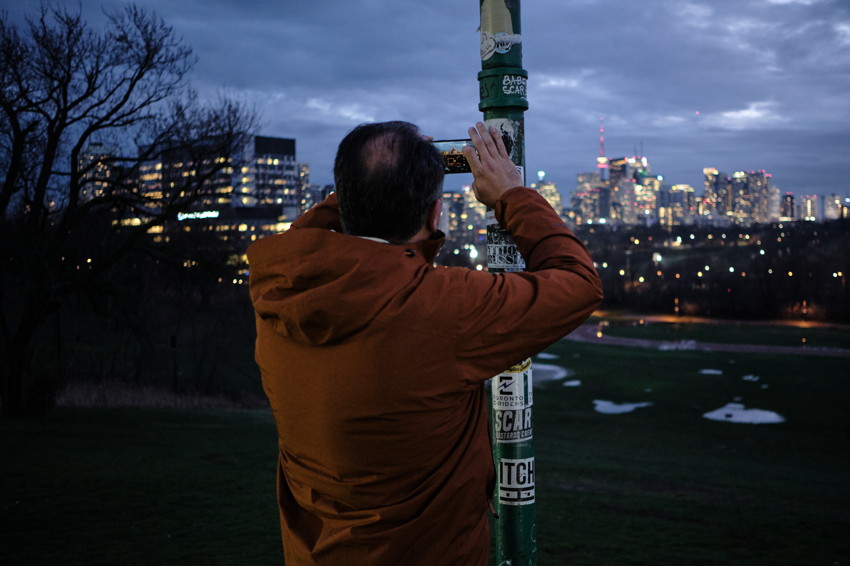 Person in orange jacket from behind, holding phone against sticker-covered pole, city skyline and park at dusk.