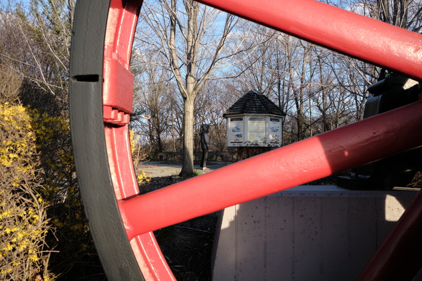 Close-up red metal wheel spokes framing a park path with information kiosk, person, leafless trees, yellow shrubs.
