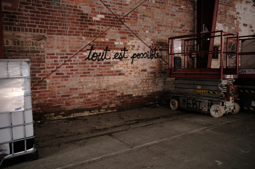 Brick wall with 'tout est possible' graffiti, red scissor lift right, plastic tank left, concrete floor.