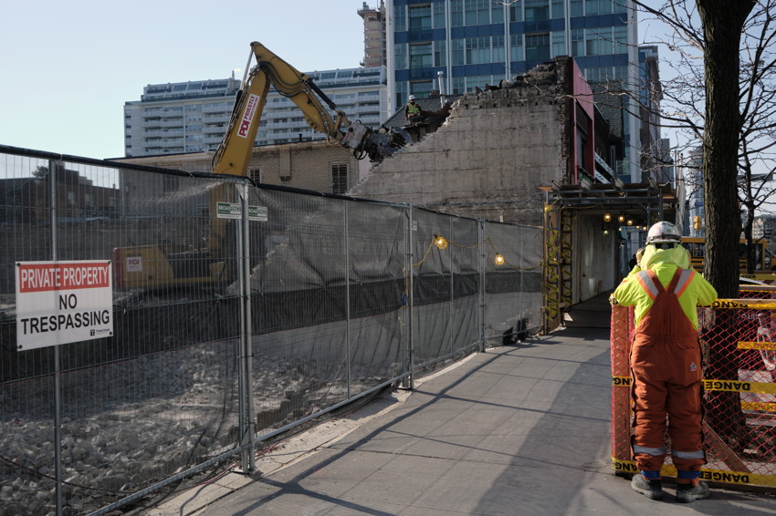 Excavator demolishing building behind fence, 'Private Property No Trespassing' sign, covered walkway with lights, worker in hi-vis.