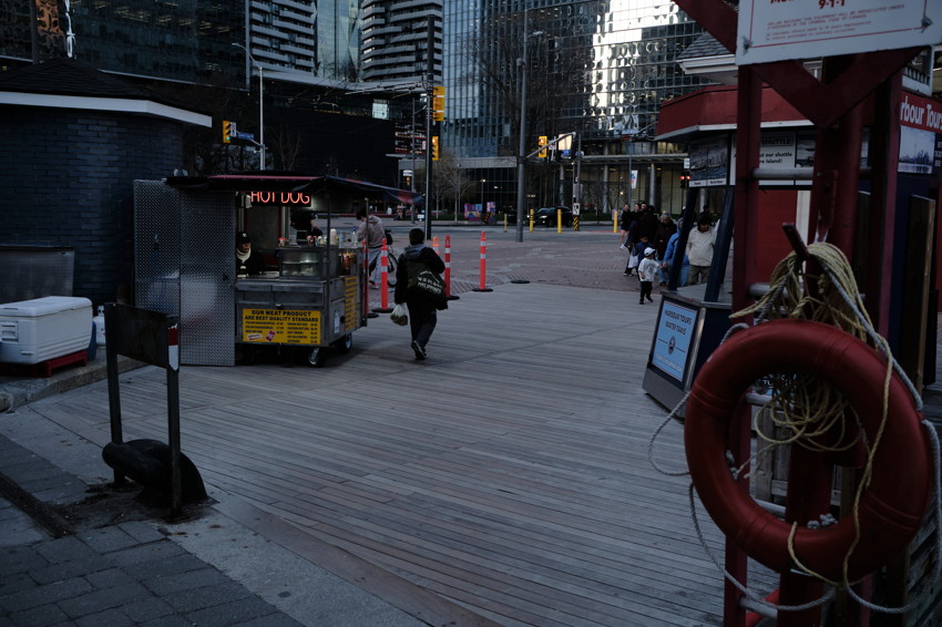 Urban boardwalk with hot dog cart left, person walking away center, life ring foreground right.