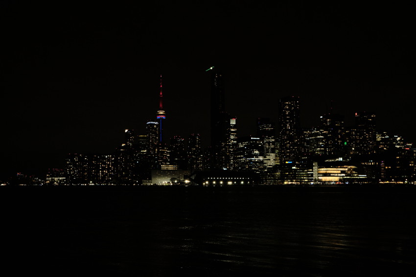Night city skyline across water with illuminated CN Tower and high-rise buildings, dark sky, light reflections on water.