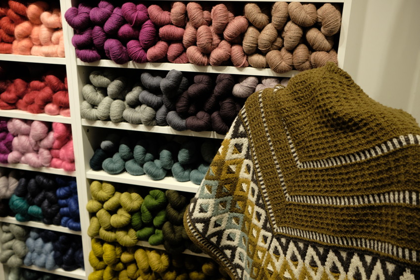 Shelves of colorful yarn skeins with an olive-green patterned knitted shawl draped in foreground.