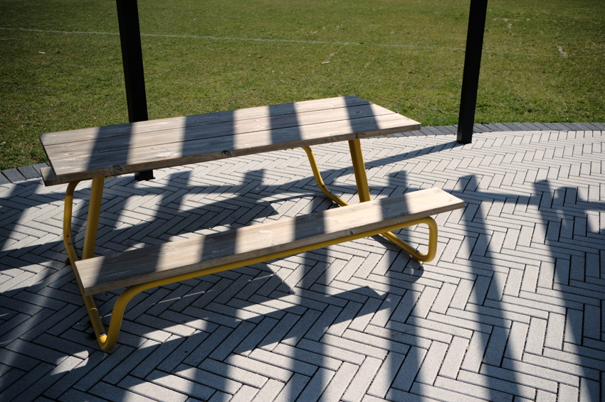 Wooden picnic table with yellow metal legs on herringbone tiled patio, long shadows from overhead structure.