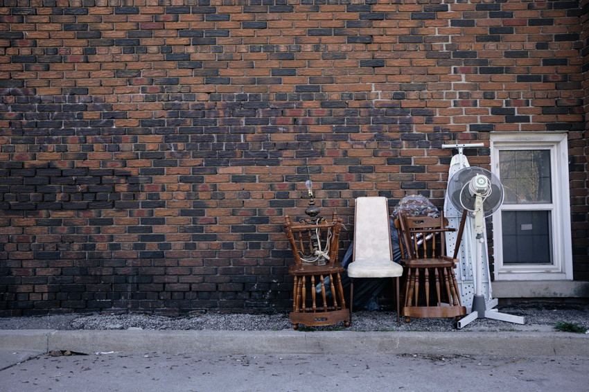 Discarded chairs, lamp, ironing board, and standing fan against a weathered brick wall beside a small window.