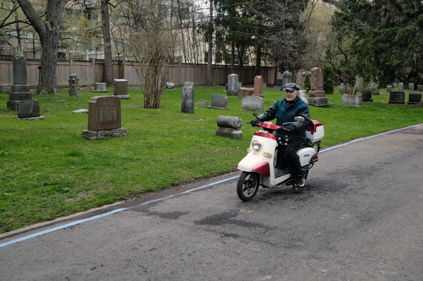 Person riding white-and-red scooter on paved path beside grassy cemetery with scattered headstones and trees.