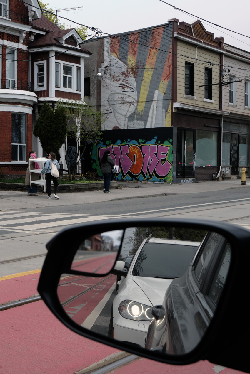 Car side mirror reflecting a car; street with graffiti mural on brick rowhouse, tram tracks, three pedestrians.