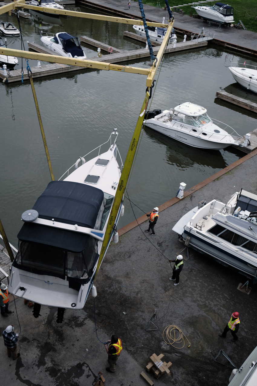 A boat suspended by slings over a marina dock while workers in safety vests guide it; nearby moored boats.