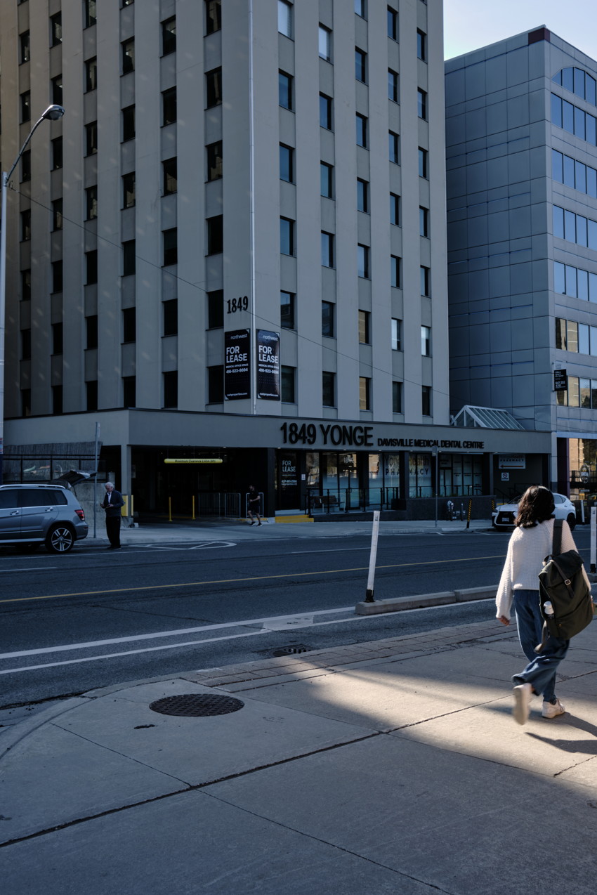 Morning light cuts diagonally across the sidewalk in front of office building as a pedestrian enters the bright strip.