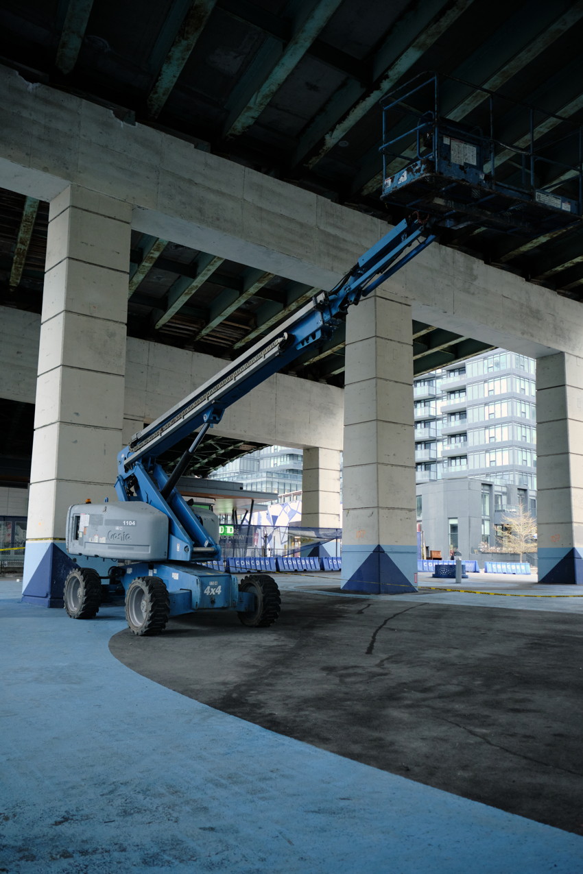 Blue articulated boom lift beneath concrete overpass, arm extended toward ceiling, large square columns, urban buildings.