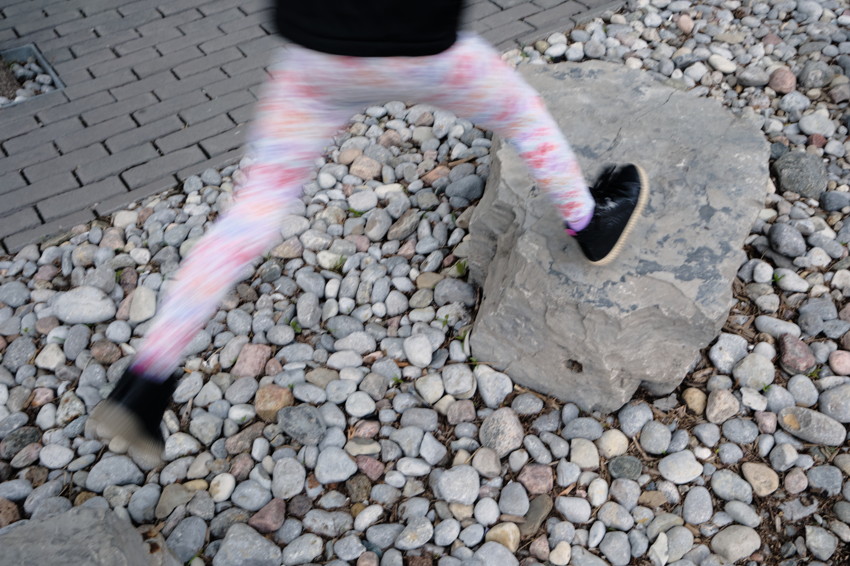 Blurred legs of a child jumping over a rock; floral leggings, black shoes, pebble ground and brick walkway.