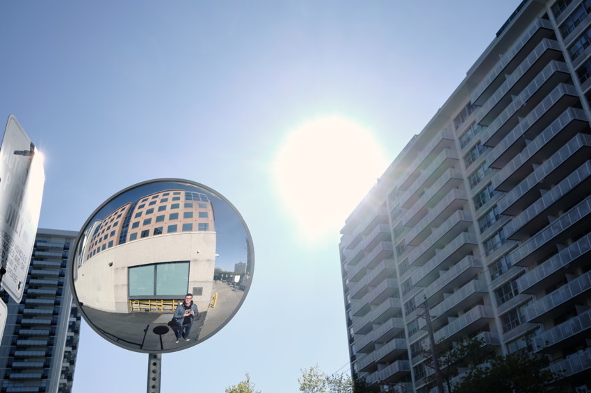 Convex street mirror reflecting a person and a building facade; adjacent high-rise apartments, bright sun, clear blue sky.