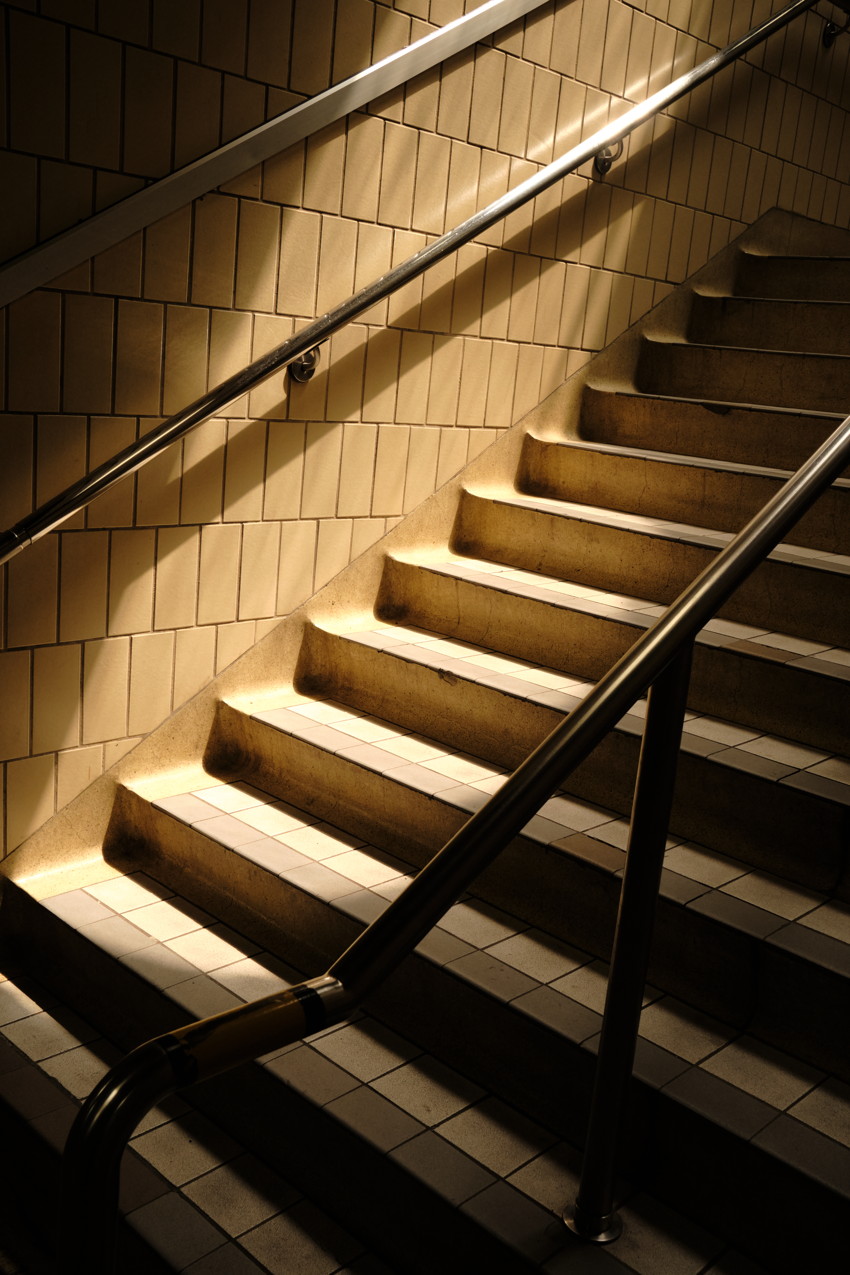 Concrete stairway with wall and metal handrails, warm diagonal light and shadow across steps.