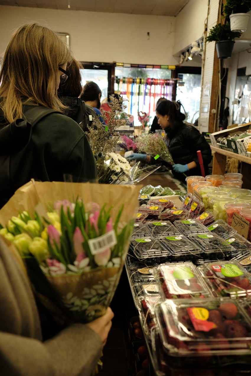 A hand holds wrapped pink and yellow tulips at a flower shop counter with packaged berries and staff arranging bouquets in the background.