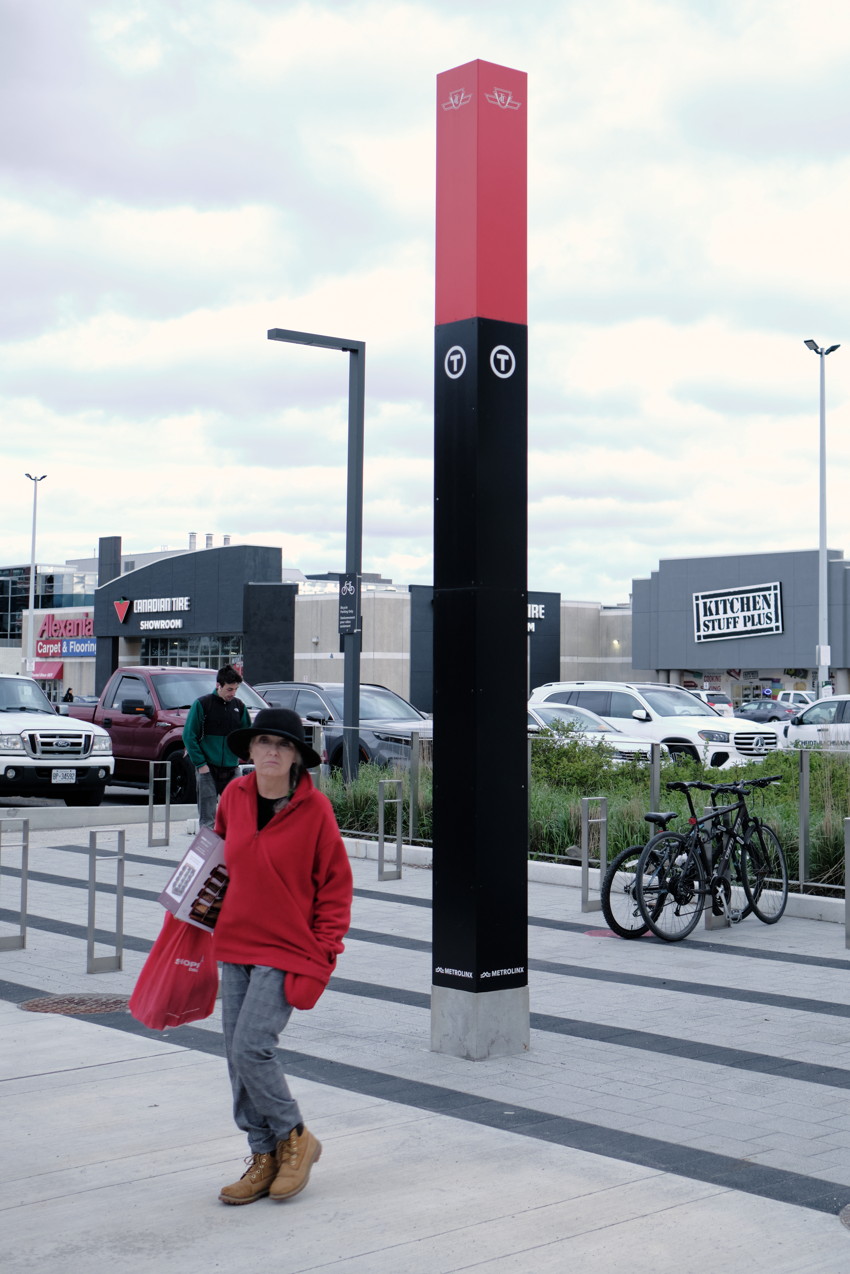 Pedestrian in red hoodie walking past tall red-and-black transit column outside shopping centre; parked cars and bicycles in background.