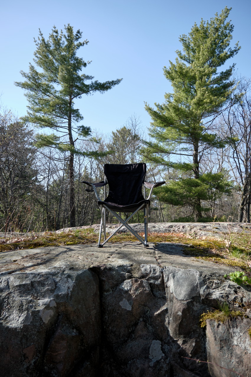 Black folding camp chair centered on a rocky outcrop, pine trees and clear sky in the background.