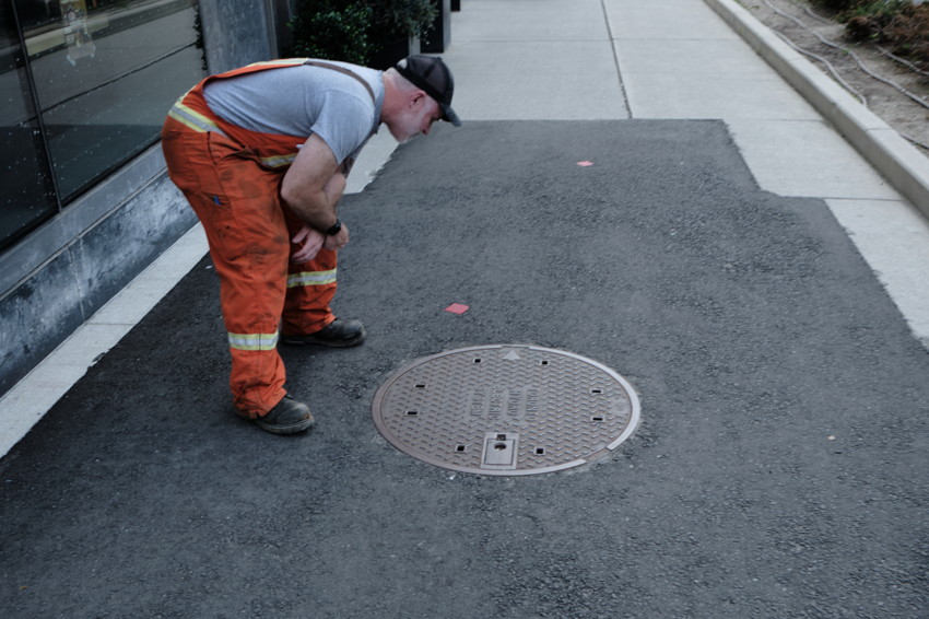 Construction worker in orange pants leaning over a round manhole cover on newly paved sidewalk.