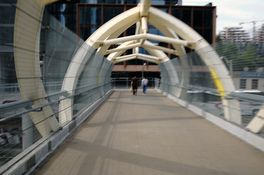 Pedestrian covered bridge with pale yellow arched beams and wire-mesh sides; two people walking away toward building.