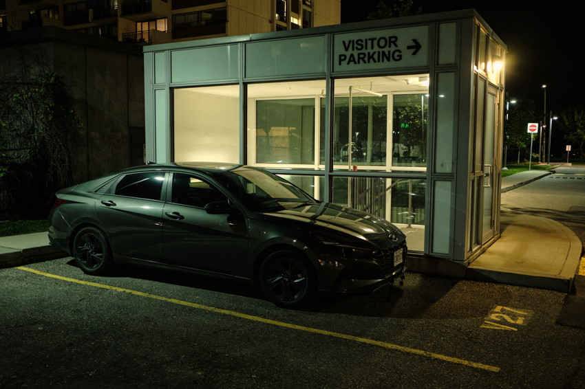 Dark sedan parked at night beside lit glass visitor parking booth; apartment building and streetlights in background.