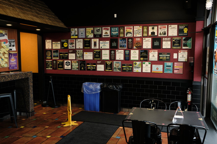 Dining area with wall of framed awards, tiled floor, table and chairs, yellow caution sign and trash bins.
