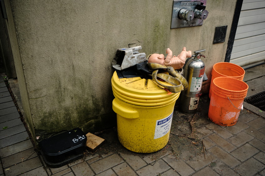 Yellow plastic bucket with lid, doll torso, fire extinguisher, two orange buckets against stained concrete wall.