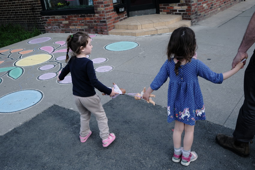 Two young girls holding dolls and an adult's hand, walking past sidewalk chalk flower in front of brick storefront.