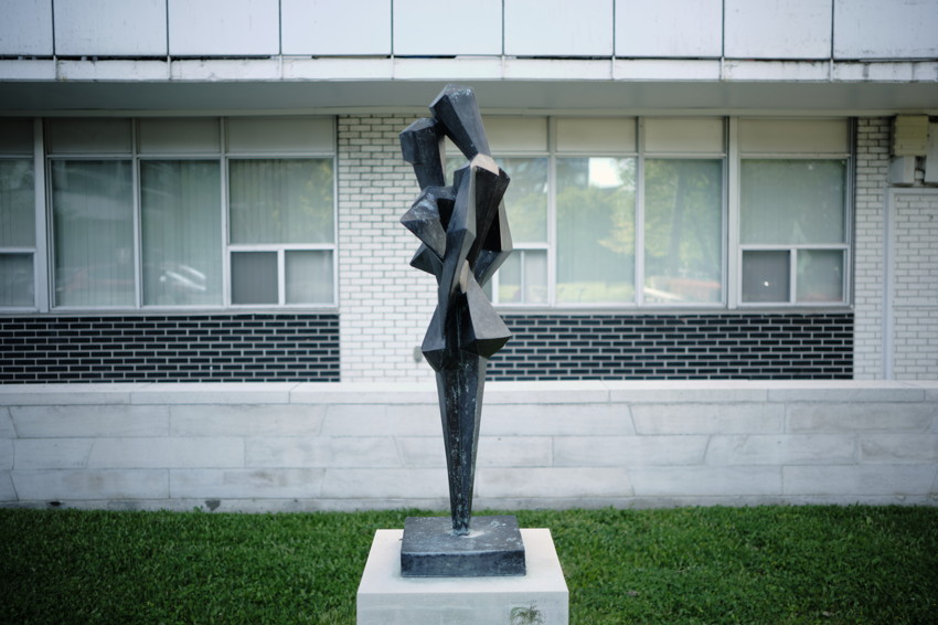Tall angular abstract dark metal sculpture on square pedestal, centered on lawn in front of mid-century building windows.