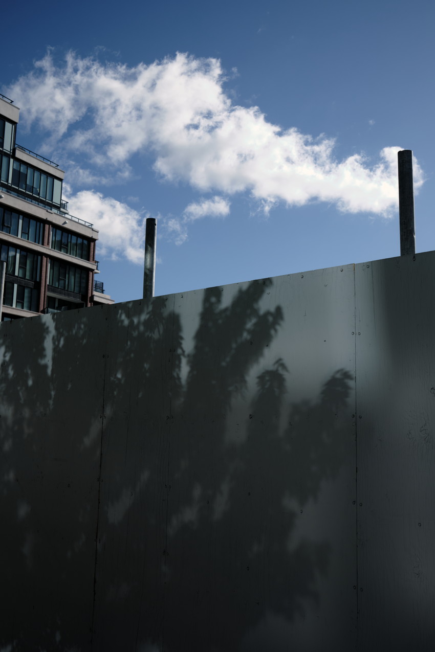 Plywood hoarding with tree shadows in foreground, apartment building left, blue sky and cloud above.