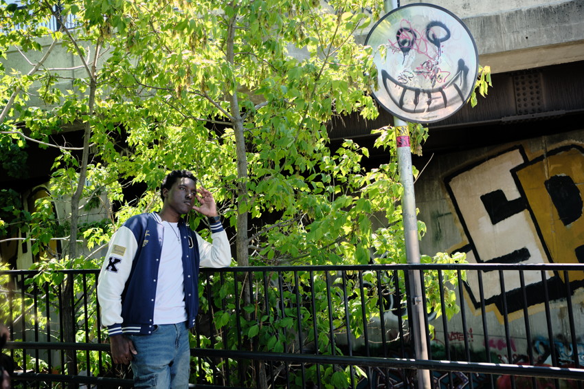Young man in varsity jacket standing by metal railing under trees, graffiti-covered wall and painted circular sign above
