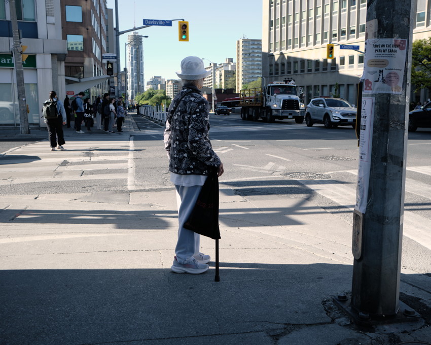 Elderly person with white bucket hat and cane stands at crosswalk facing street, trucks and pedestrians behind.