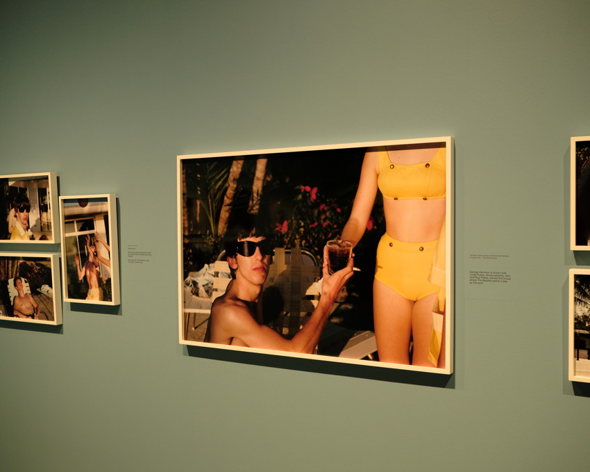 Gallery wall showing framed photo: shirtless person in sunglasses raising drink toward standing person in yellow bikini.